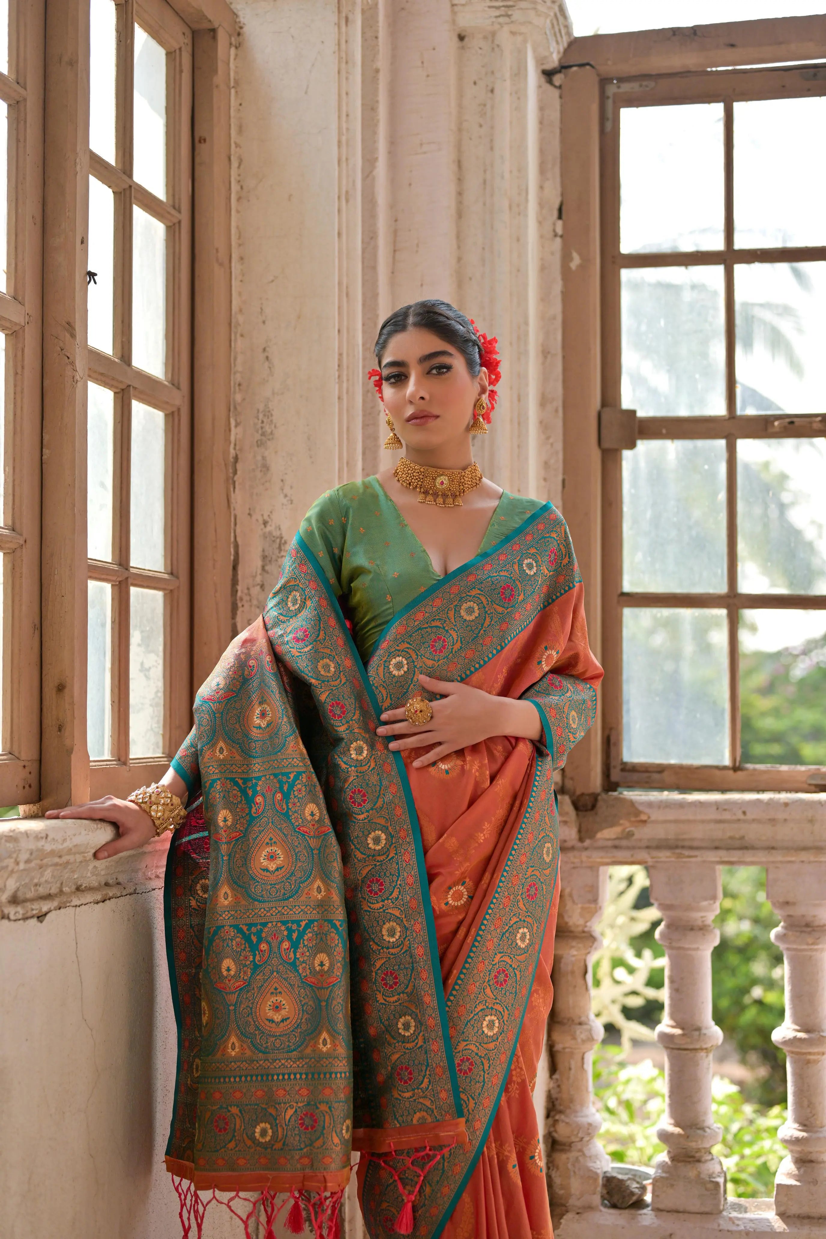 A woman models a vibrant Coral Orange Banarasi Silk Saree with all-over gold Zari weaving. The saree has a wide, ornate Rama Blue/Teal border featuring traditional Zari weaving. She wears a light green blouse and heavy gold Kundan jewelry, posing by a large antique window.